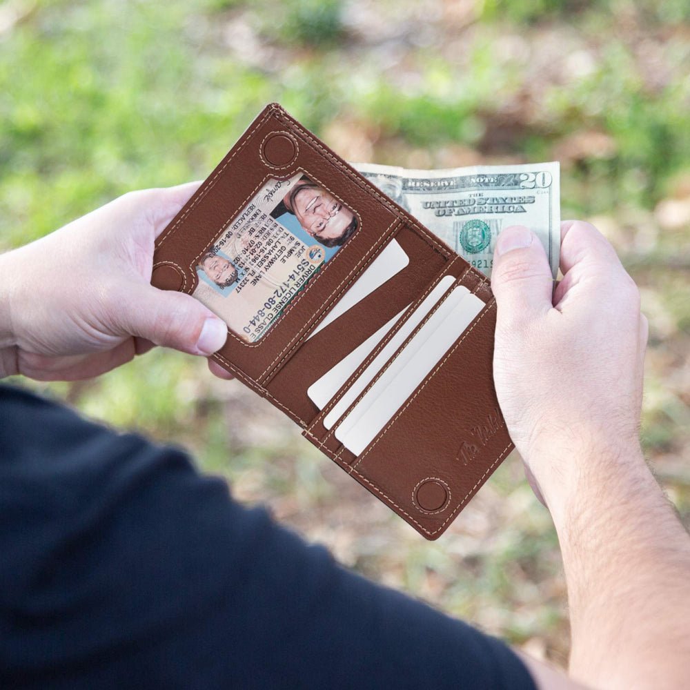 Brown leather wallet with card slots and a small pocket, held by hands against a blurred green outdoor background.